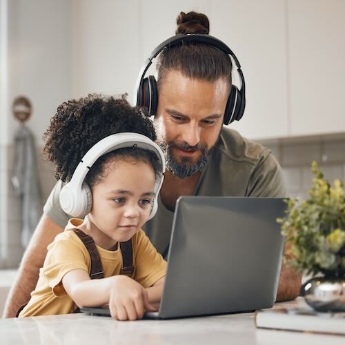 Ein-gluecklicher-Vater-sitzt-mit-seiner-jungen-Tochter-am-Kuechentisch-beide-haben-kopfhoerer-auf-und-blicken-in-den-vor-sich-aufgeklappten-laptop-und-hoeren-einen-spirituelle-podcast